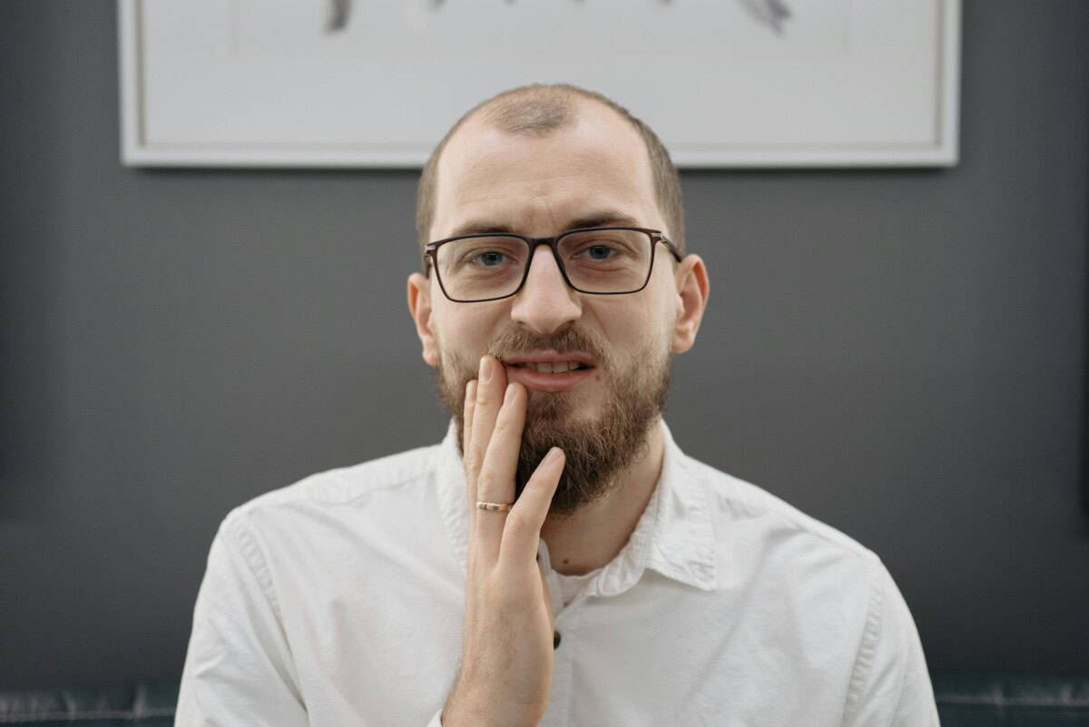 A man with eyeglasses and beard shows discomfort from a toothache indoors.